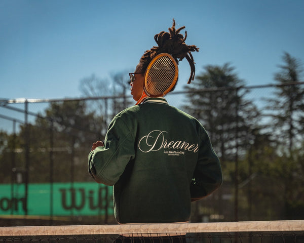 Person holding a tennis racket on a tennis court with 'Dreamer' jacket